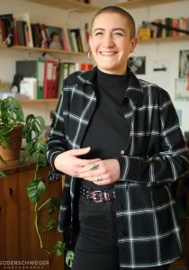 Portrait von Rosa Jellinek, CO-Vorsitzende von Keshet Deutschland e.V. Rosa Jellinek steht neben einem Regal mit einer Zimmerpflanze. Sie lächelt in die Kamera.